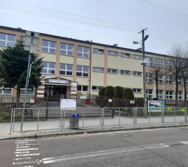 Na zewnątrz, pochmurny dzień. Zdjęcie szkoły przed którą stoją latarnie z zamontowanym monitoringiem