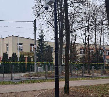 Na zewnątrz, pochmurny dzień. Zdjęcie alejki przy której stoi latarnia z zamontowanym monitoringiem, w tle ogrodzenie i...