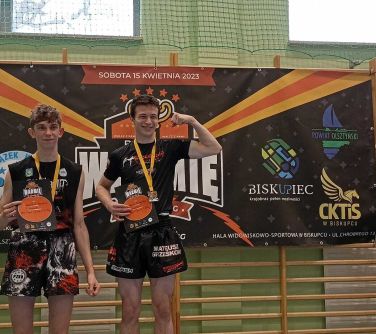 dwóch nastolatkó pozuje na podium, w tle baner, po prawej stronie baner patronat starosty olsztyńskiego