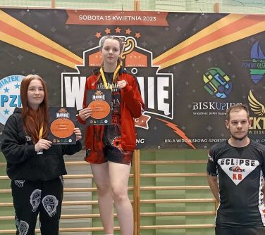 dwie nastolatki pozują na podium, w tle baner, po prawej stronie stoi mężczyzna z rękami z tyłu