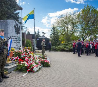 poczet honorowy przy pomniku usłanym kwiatami, po lewej stronie stoi orkiestra dęta i gra, po środku przed czarnym namiotem...