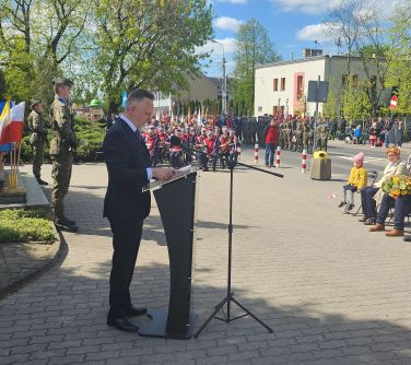 mężczyzna przemawia przy mównicy, w tle żołnierze przy pomniku i siedząca orkiestra dęta