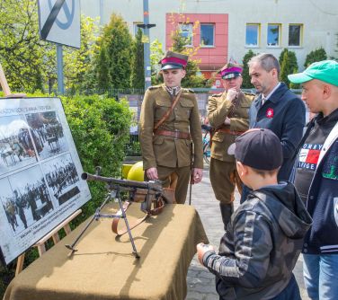 osoby oglądaja karabin przed którym stoi obraz ze starymi zdjęciami