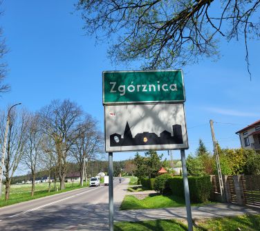 zdjęcie tablicy z napisem miasta Zgórznica, w tle ulica, samochody po prawej stronie dom z ogrodzeniem