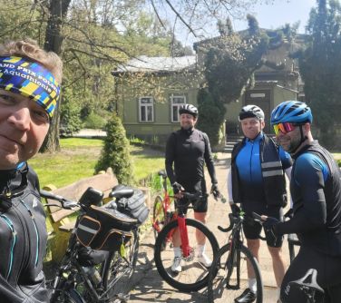czterech rowerzystów stoi przy rowerach na chodników, obok drewniana ławka, w tle domek