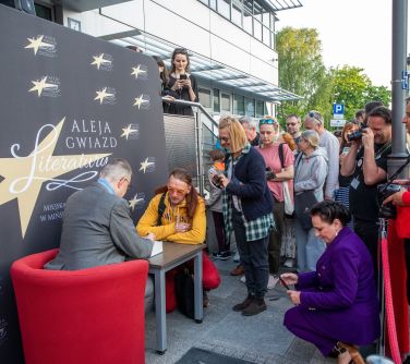 mężczyzna siedzi w fotelu przy stoliku składa autografy zgromadzonym osobom