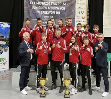 zawodnicy z trenerem pozują do zdjęcia, część stoi z tyłu na podium, reszta przed podium, po obu stronach mężczyźni w...