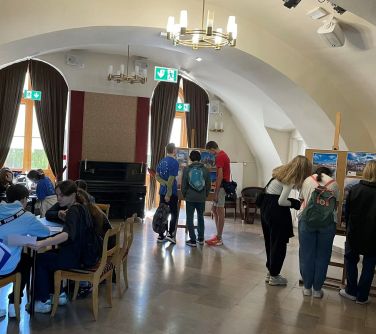 wnętrze domu kultury osoby siedzą przy stole, inni oglądają teksty wywieszone na sztaludze