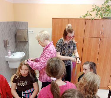 dziewczyni, dwie siedza na krzesłach nad nimi dwie panie plotą warkocze na włosach