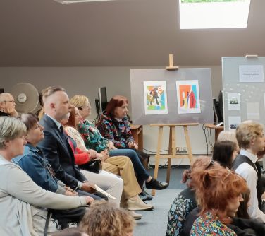 osoby siedza na krzesłąch słuchają w tle prace na stelarzach