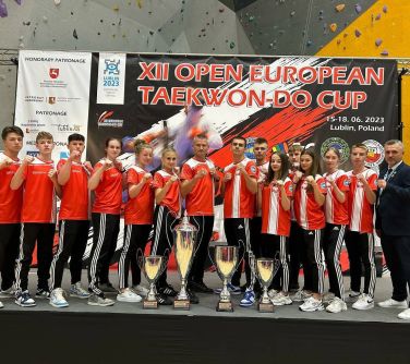 grupa osób pozuje do zdjęcia, przed nimi cztery puchary różnej wielkości, w tle baner XII Open European Taekwo-Do Cup