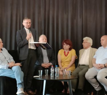 mężczyżmi i kobieta siedzą przy stoliku na scenie, mężczyzna w koloratce mówi do mikrofonu trzymając książkę