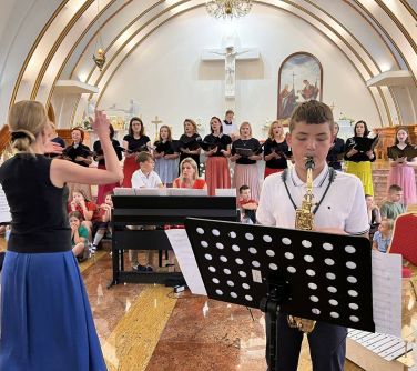 wnętrze kościoła koncert chóru, tyłem stoi kobieta dyryguje, przodem chłopiec gra na trąbce przed nim pulpit z nutami