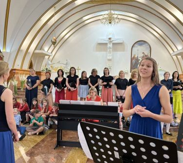wnętrze kościoła wystep chóru po lewej stronie tyłem stoi kobieta, po prawej przodem kobieta śpiewa przed nią stojak na nuty