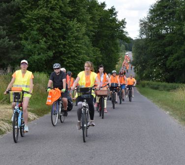 uczestnicu rajdu rowerowego w pomarańczowych kamizelkach jadą na rowerach wiejską drogą asfaltową, na pierwszym planie dwie...