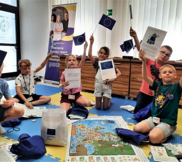 grupa dzieci siedzi na piankowych matach unosi ręce do góry trzymając flagi ue i czasopisma, na matach worki, plansze