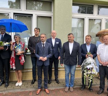 grupa ludzi stojących pod buydynkiem z lewej strony kobieta trzyma znicz, mężczyzna bukiet kwiatów, stoją pod parasolem