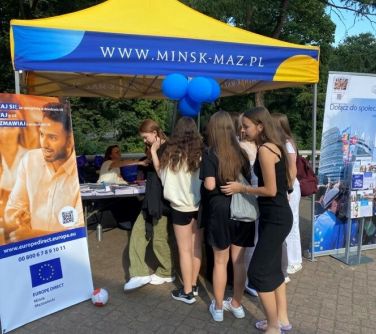 niebiesko-żółty namiot pod którym stoi grupa dziewcząt, oglądająca publikacje rozłożone na stole