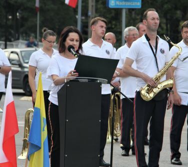 Kobieta stoi przy mównicy i wygłasza przemówienie, w tle orkiestra dęta