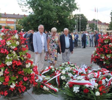 dwóch mężczyzn i kobieta z wieńcem stoją przy pomniku niepodległości