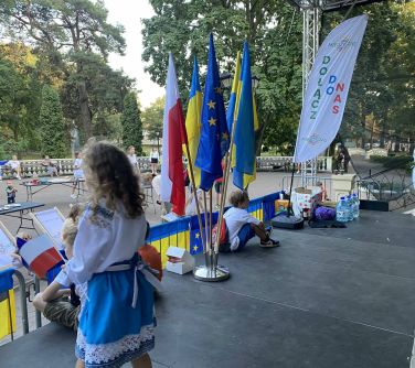 dzieci stoją i siedzą na scenie, na przodzie sceny flagi, poniżej sceny stoły z jedzeniem i gadżetami pomiędzy przechadzają...