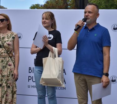 trzy osoby stoją na scene, mężczyzna przemawia do mikrofonu