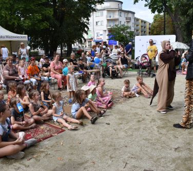 grupa osób siedzi na dywanach i krzesłach słucha przedstawienia