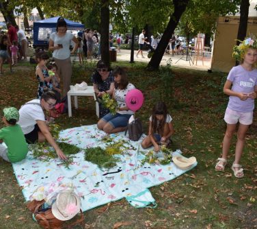 grupa osób siedzi na kocu i plotą wianki z kwiatów polnych, obok stoi dziewczynka z wiankiem na włosach, w tle osoby na...