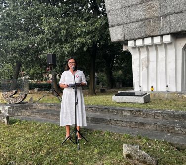 kobieta przemawia do mikrofonu, trzyma w dłoniach kartki, w tle drzewa i postument