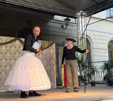 kobieta i mężczyzna na scenie, mężczyzna po lewej w spódnicy baleriny, z wachlarzem w dłoni, po prawej stronie kobieta z...