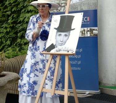 kobieta w kapeluszu stoi obok palety ze zdjęciem, opowiada, w tle baner obok krzew