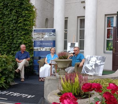 trzy osoby siedzą na fotelach na wejściu do muzeum za nimi dwa filary, po lewej stronie filarów baner, po prawej stronie...