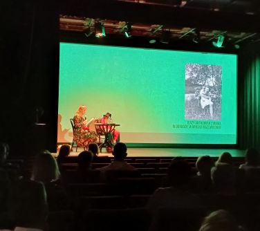 dwie osoby siedzą na scenie na krzesłach przy stoliczku, w tle wyświetlane stare zdjęcie, publiczność siedzi na widowni