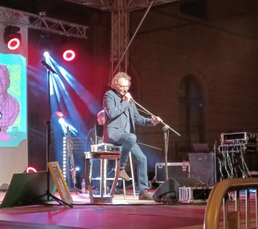 mężczyzna siedzi na krześle na scenie przed nim mikrofon do którego mówi, w tle światła