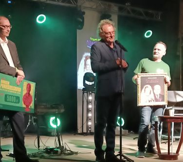 trzech mężczyzn stoi na scenie, od lewej mężczyzna trzyma duży czek na kwotę 5000 zł, mężczyzna po środku stoi przed...