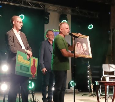 trzech mężczyzn na scenie, na przodzie mężczyzna trzyma obraz i przemawia