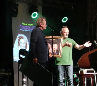 dwóch mężczyzn stoi na scenie, mężczyzna po prawej stronie wskazuje w kierunku publiczności, po lewej stronie mężczyzna...