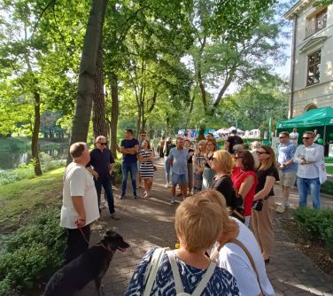 grupa osób słucha przewodnika stojąc na alejce w parku , w tle drzewa i woda