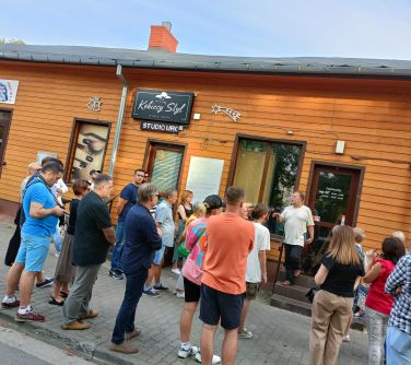 grupa osób słucha przewodnika stojąc na chodniku przed drewnianym domkiem