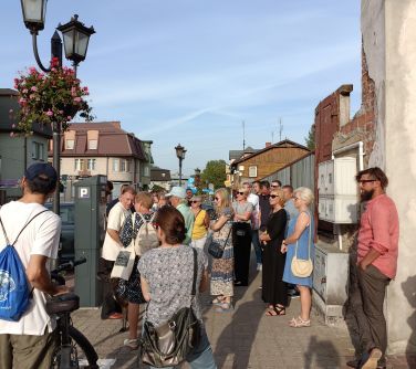 grupa osób słucha przewodnika stojąc na chodniku między domami