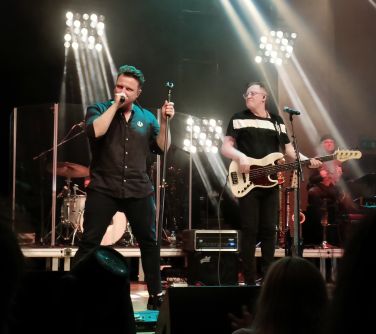 mężczyzna śpiewa na scenie, po prawej stronie mężczyzna gra na gitarze