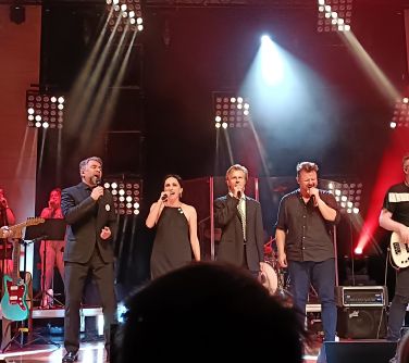 czterech wykonawców śpiewa na scenie, po prawej i po lewej stronie mężczyzni grają na gitarze