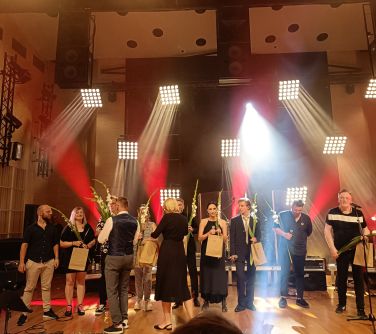 osoby/wykonawcy stoją na scenie, mężczyzna i kobieta wręczaja im kwiaty i upominki