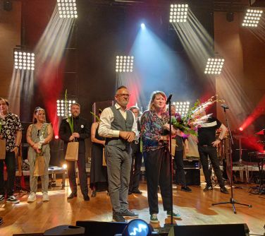 kobieta i mężczyzna stoją na scenie przed mikrofonem, kobieta trzyma w dłoniach kwiaty, za nimi stoją wykonawcy z kwiatami...