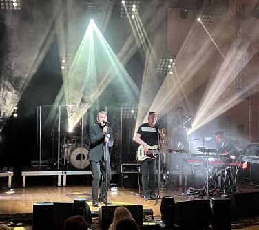 mężczyzna śpiewa na scenie, po prawej stronie mężczyzna gra na gitarze, drugi na klawiszach, trzeci na trąbce, w tle...