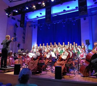 na scenie z tyłu chór stoi, z przodu orkiestra siedzi bokiem na krzesłach gra na instrumentach, po środku na przodzie na...