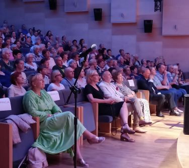 publicznośc na fotelach słucha koncertu, przed nimi stoi mikrofon