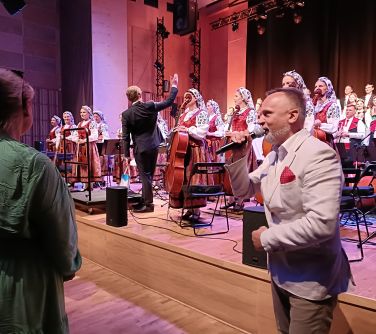 po lewej kobieta tyłem, po prawej mężćzyzna przodem przemawia w tyle na scenie chór i orkiestra, dyrygent z podniesioną...