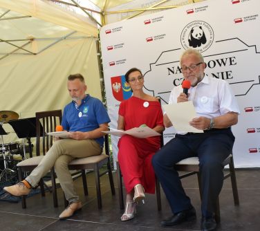 trzy osoby siedza na krzesłach na scenie pod zadaszeniem, mężczyzna po prawej stronie czyta, w tle baner narodowe czytanie