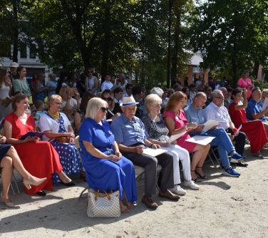 widownia siedzi na krzesłach w dwóch rzędach, za nimi osoby stoją, w tle drzewa